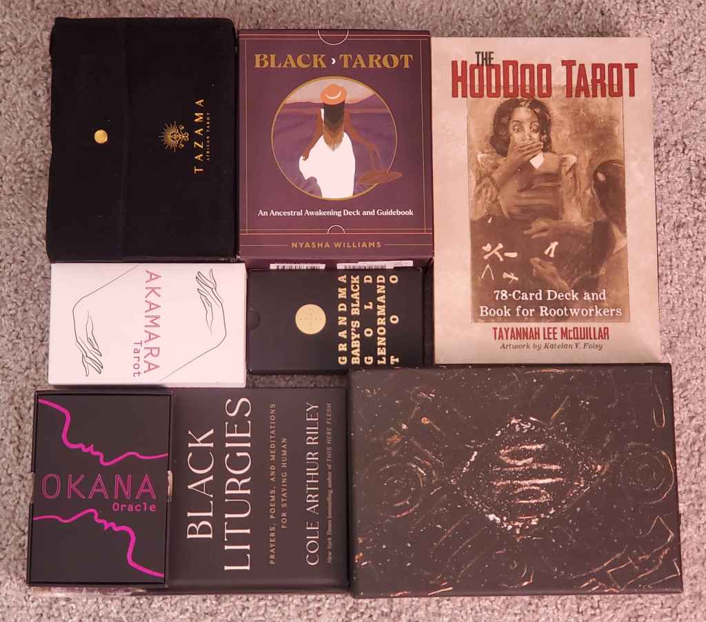 ID: several decks and books by Black creators, including Black Tarot, Okana Oracle, Akamara Tarot, Dust 2 Onyx Tarot, Grandma Baby's Black Lenormand Too, and the book Black Liturgies by Cole Arthur Riley