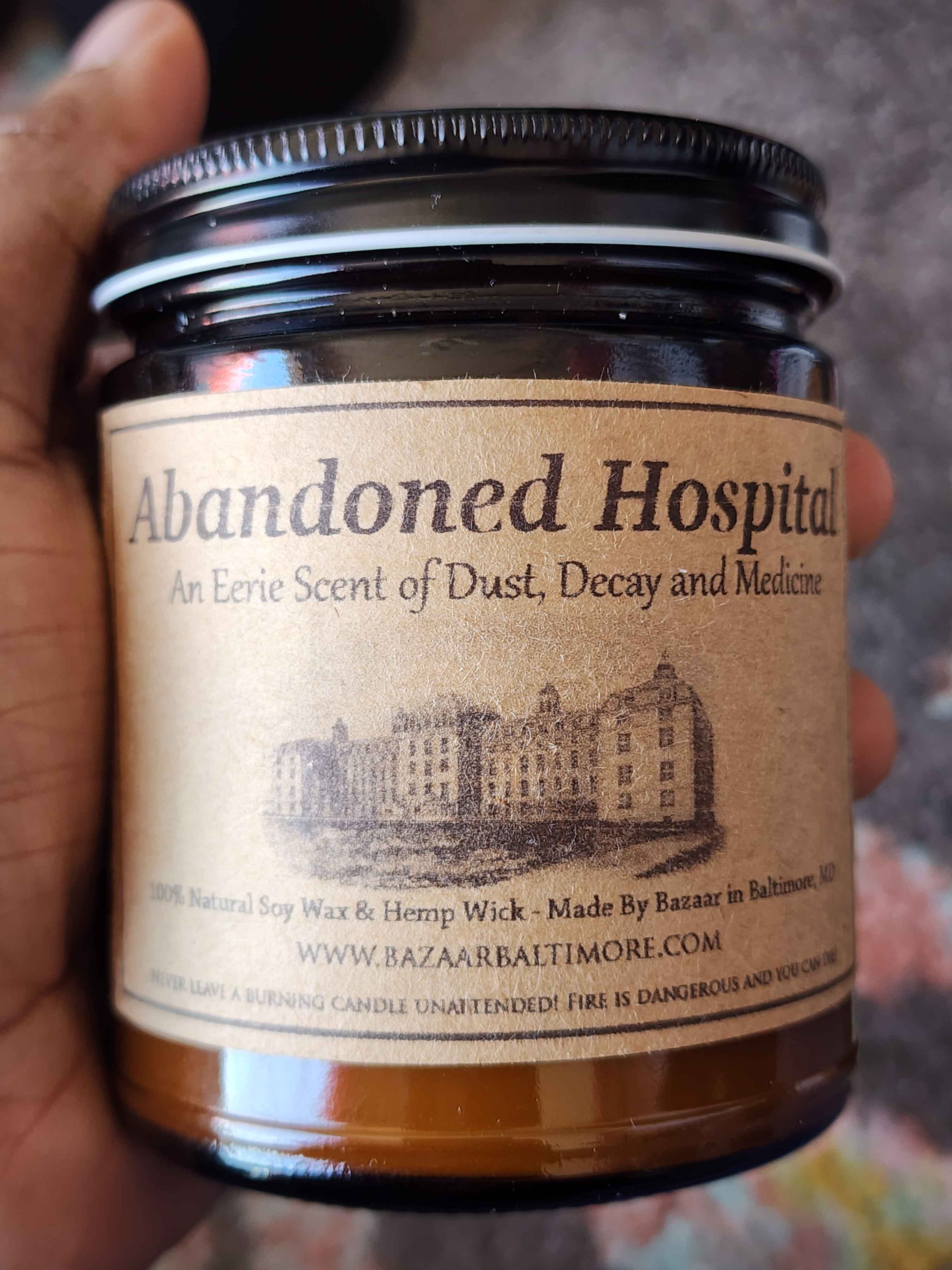 ID: an amber jar candle from Bazaar in Baltimore called Abandoned Hospital, described as an eerie scent of dust, decay, and medicine, the label includes an illustration of a 19th century hospital on it.