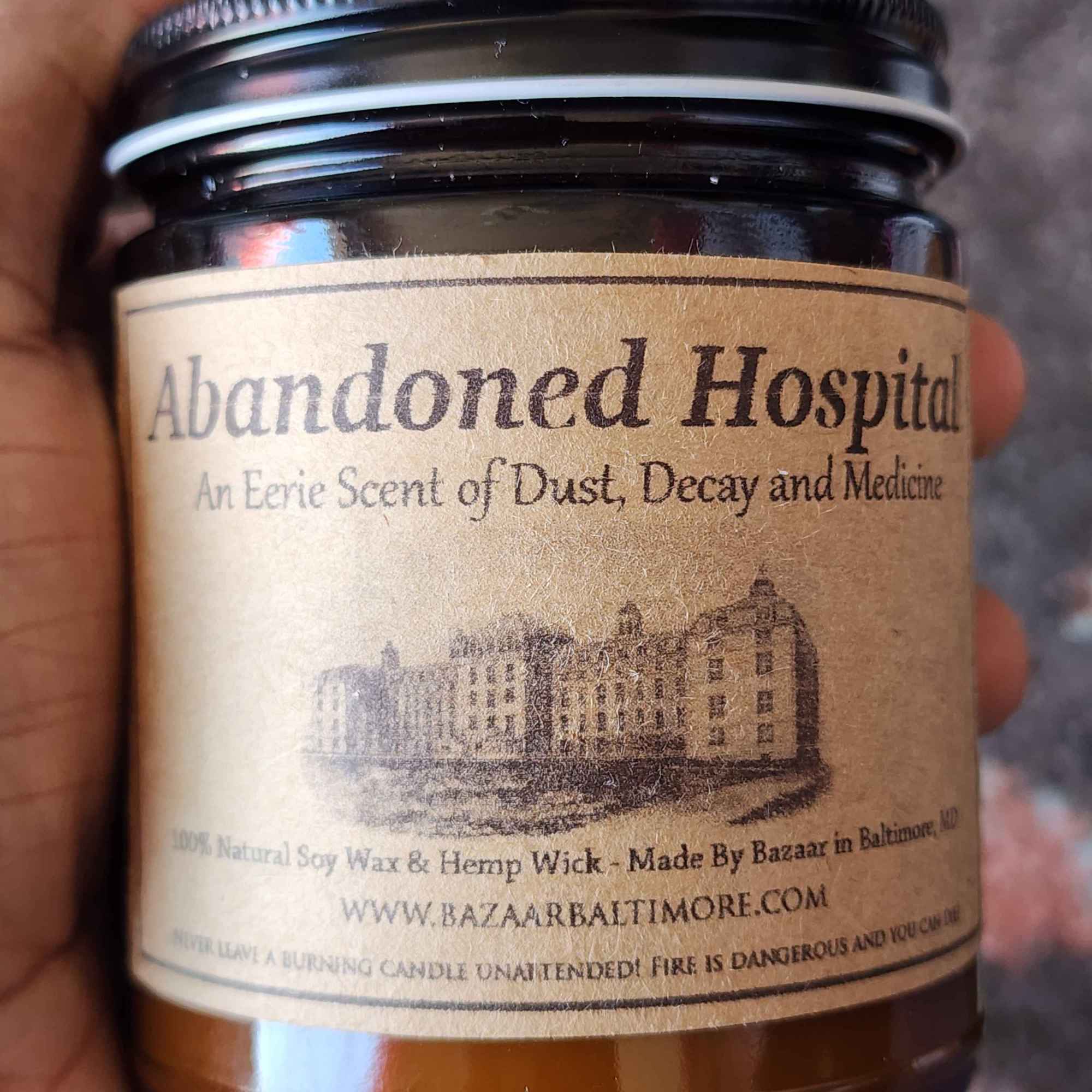 ID: an amber jar candle from Bazaar in Baltimore called Abandoned Hospital, described as an eerie scent of dust, decay, and medicine, the label includes an illustration of a 19th century hospital on it.