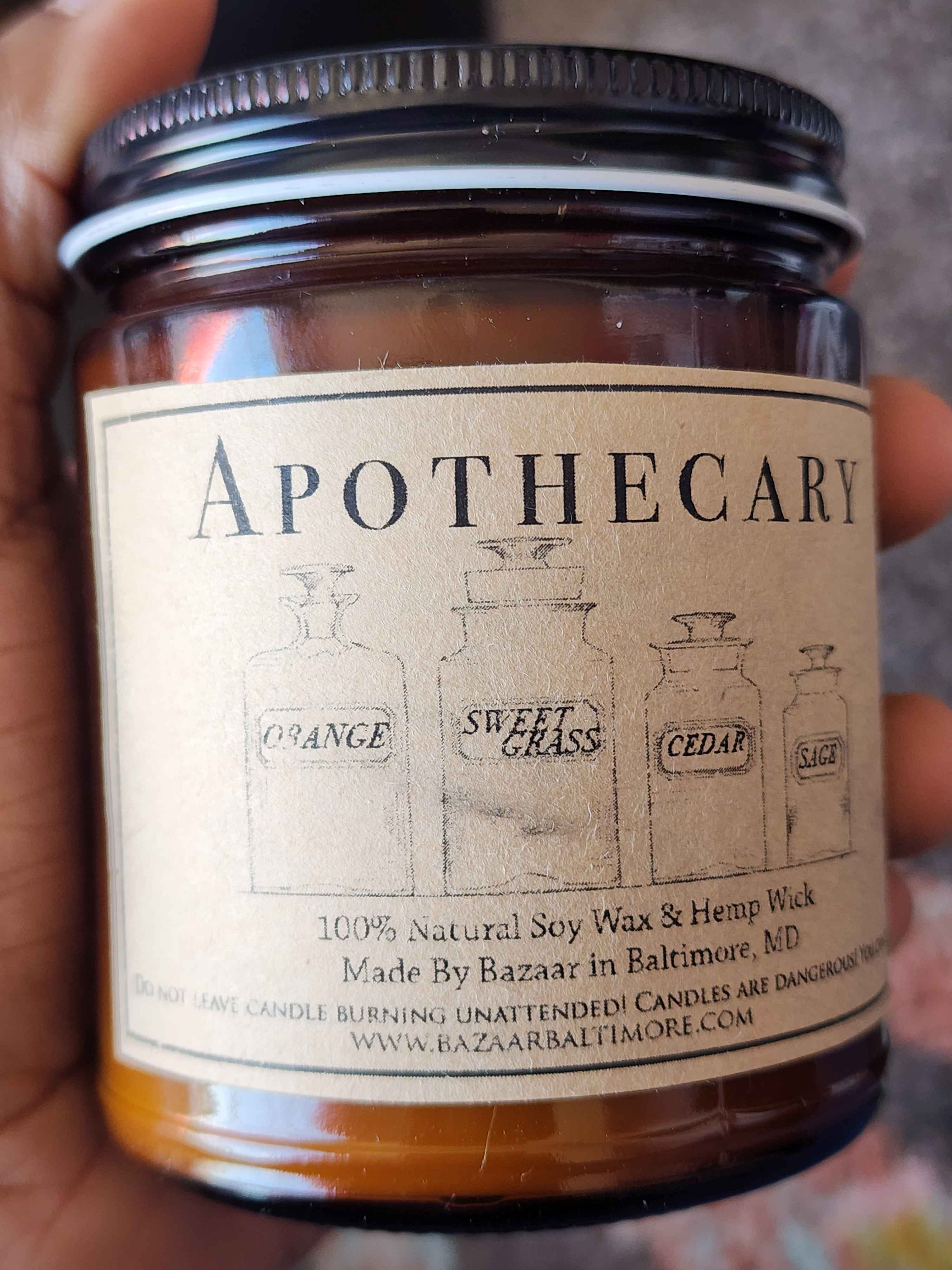 ID: an amber jar candle from Bazaar in Baltimore called apothecary. four illustrated old-time collection jars are labeled orange, sweet grass, cedar, and sage, which are the cand;e's fragrances