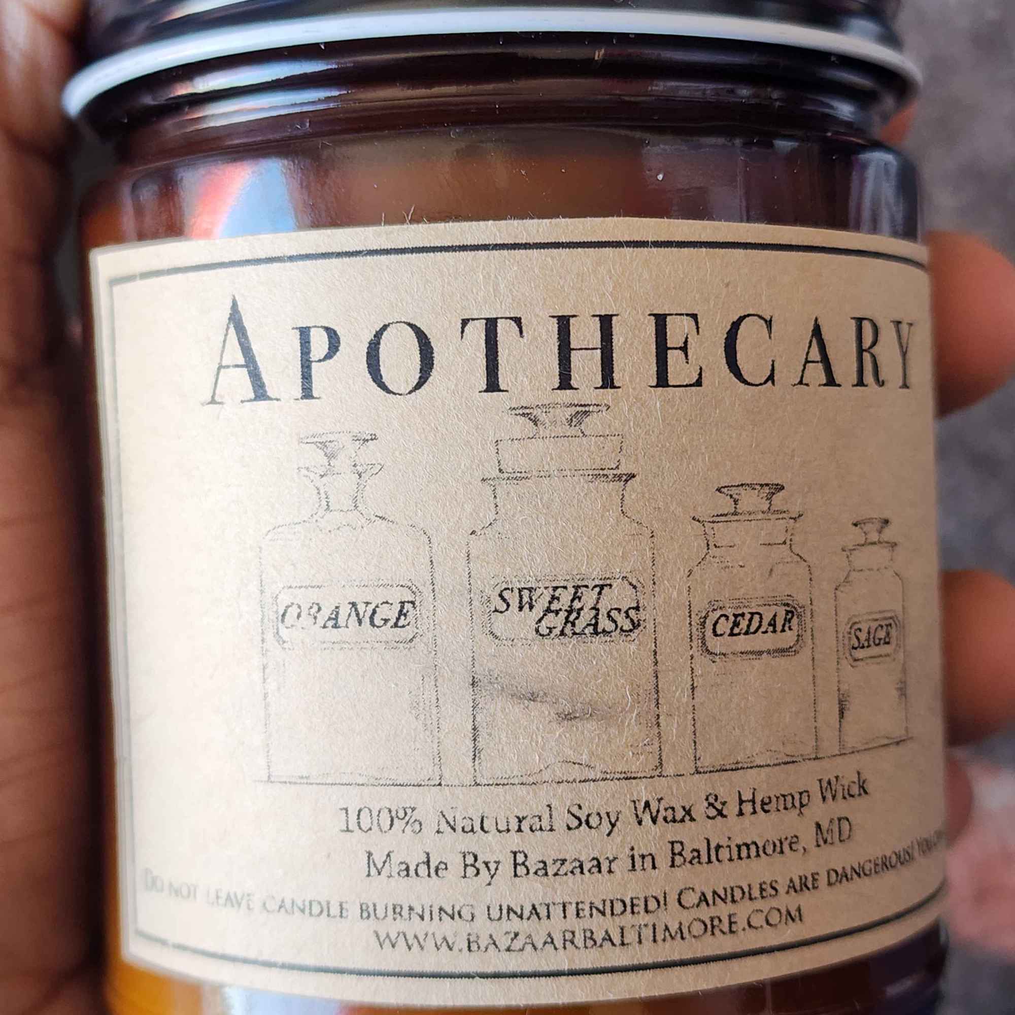ID: an amber jar candle from Bazaar in Baltimore called apothecary. four illustrated old-time collection jars are labeled orange, sweet grass, cedar, and sage, which are the cand;e's fragrances