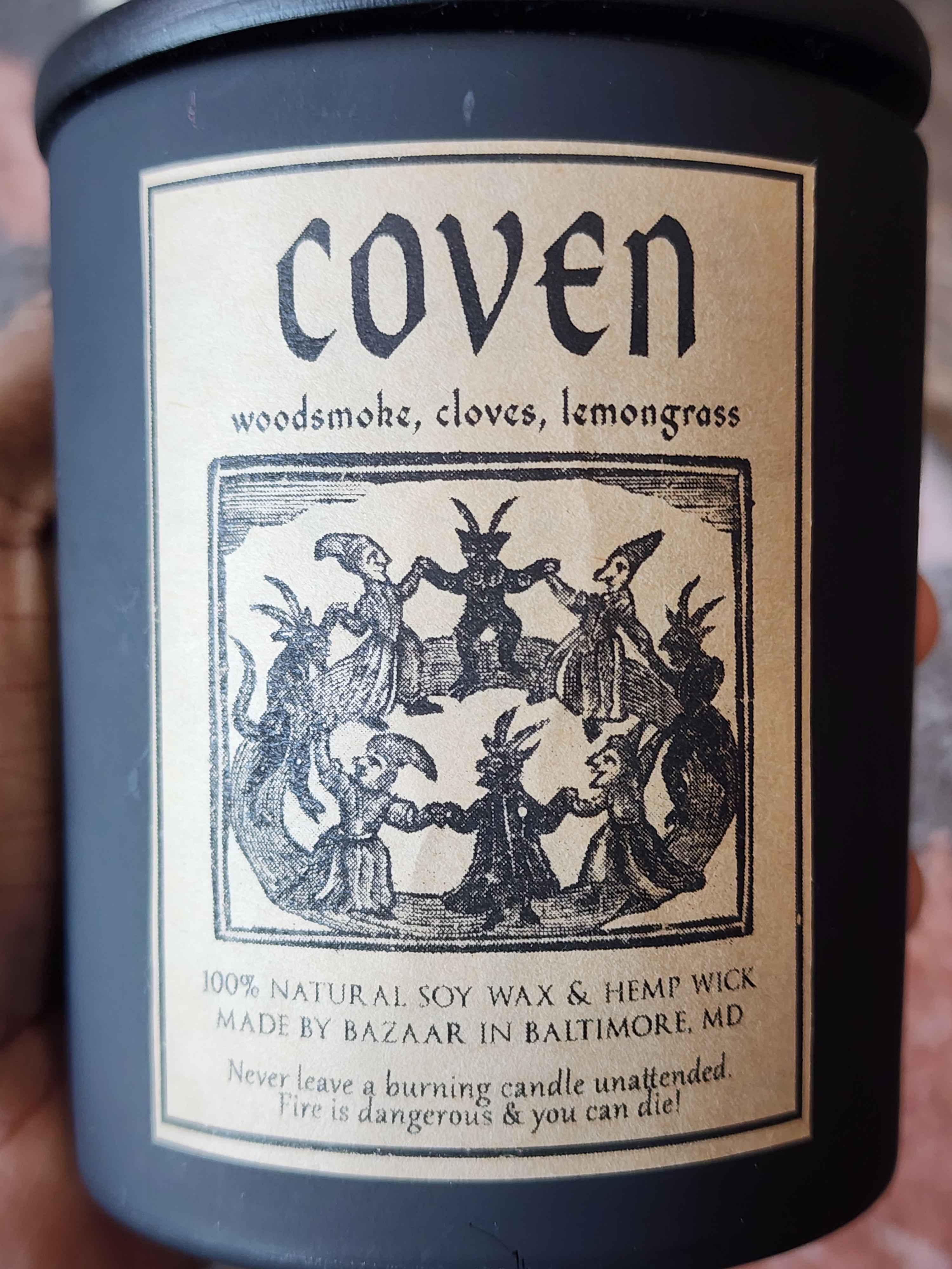 ID: a large candle in a matte black jar from Bazaar Baltimore. the candle is called Coven, and has an illustration of a witch's circle on the label. the candle has notes of woodsmoke, cloves, and lemongrass.