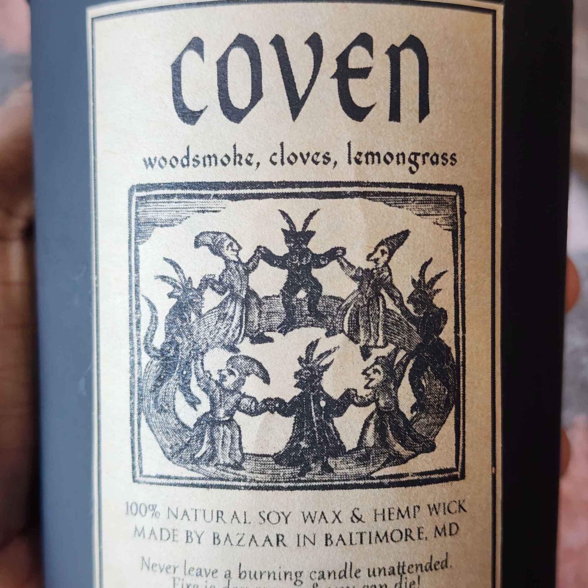 ID: a large candle in a matte black jar from Bazaar Baltimore. the candle is called Coven, and has an illustration of a witch's circle on the label. the candle has notes of woodsmoke, cloves, and lemongrass. 