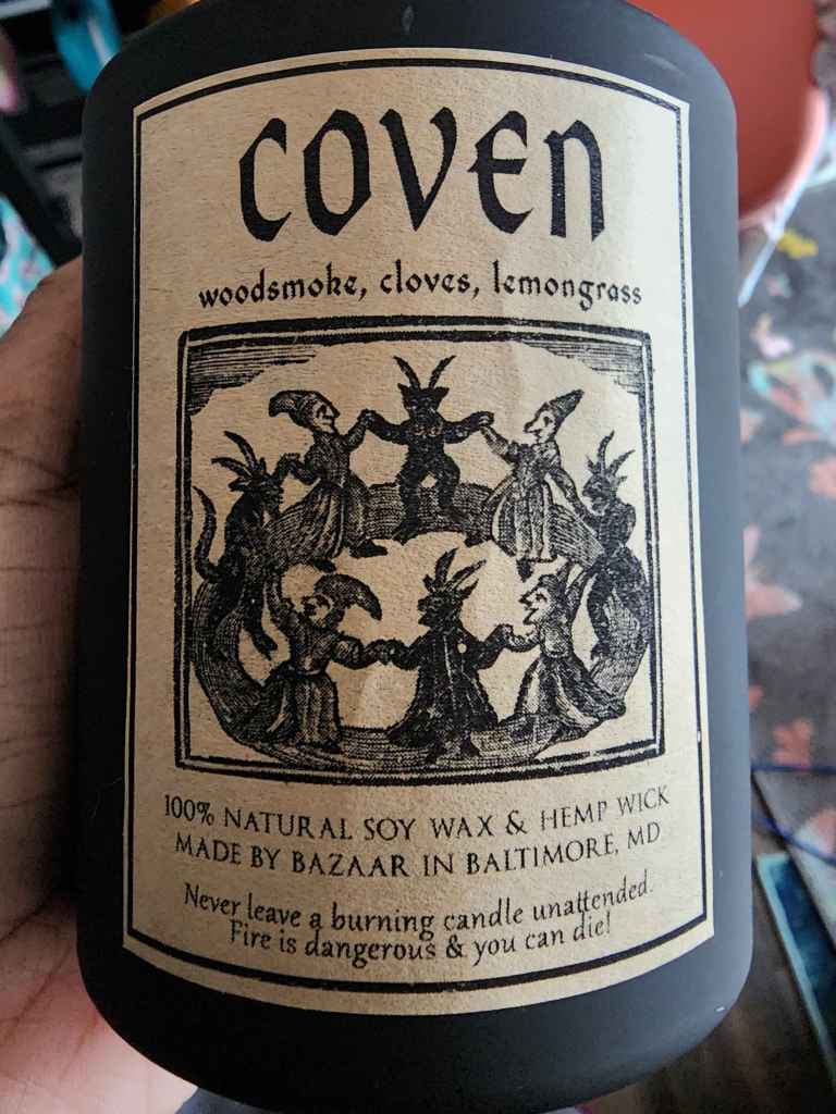 a candle in a black jar from Bazaar in Baltimore. the scent on the label is Coven, with notes of woodsmoke, cloves, and lemongrass.