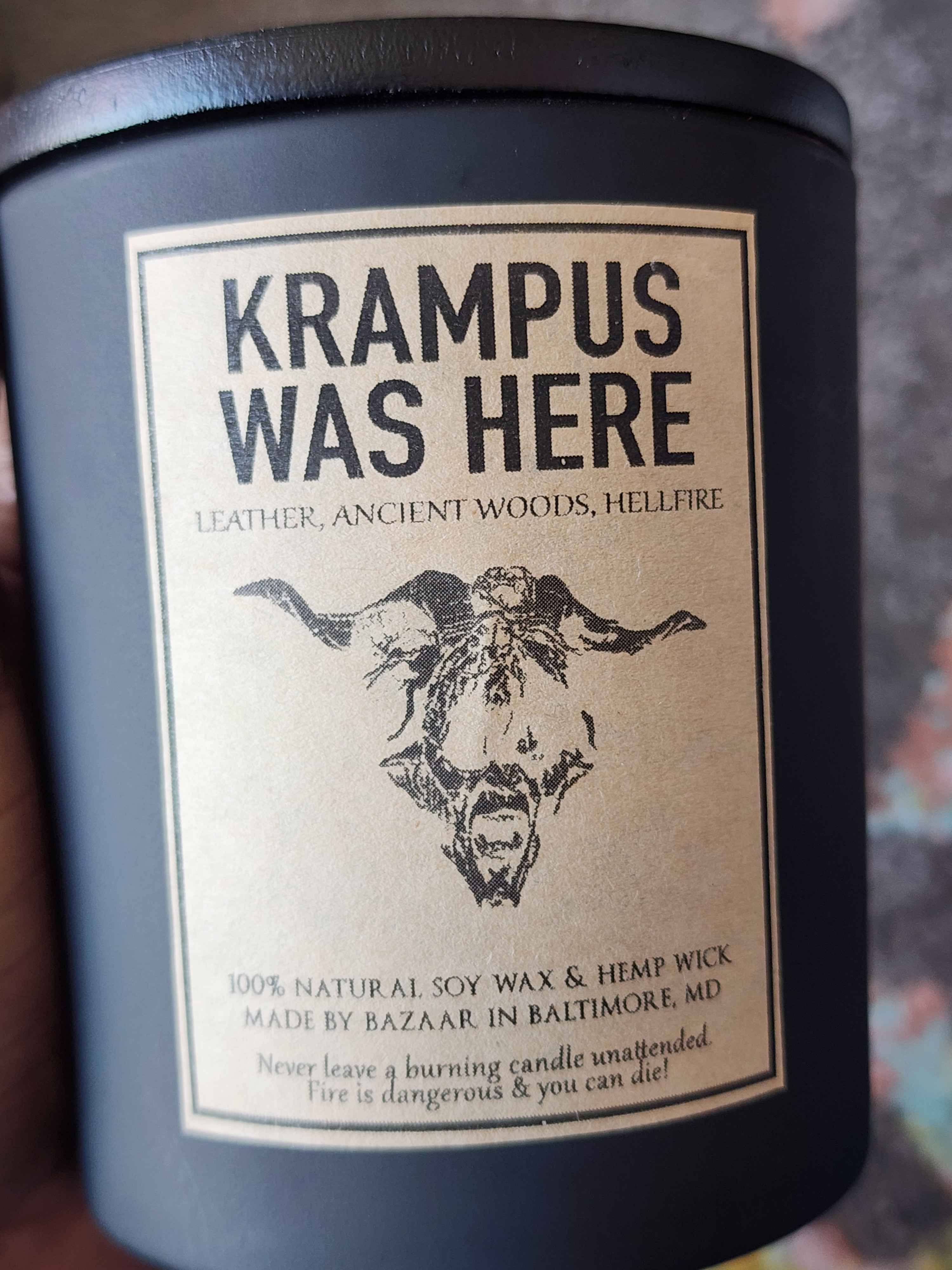 ID: a large candle from Bazaar in Baltimore in a matte black jar. the candle is called Krampus Was Here, and has an illustration of a goat head on the label. it smells like leather, ancient woods, and hellfire