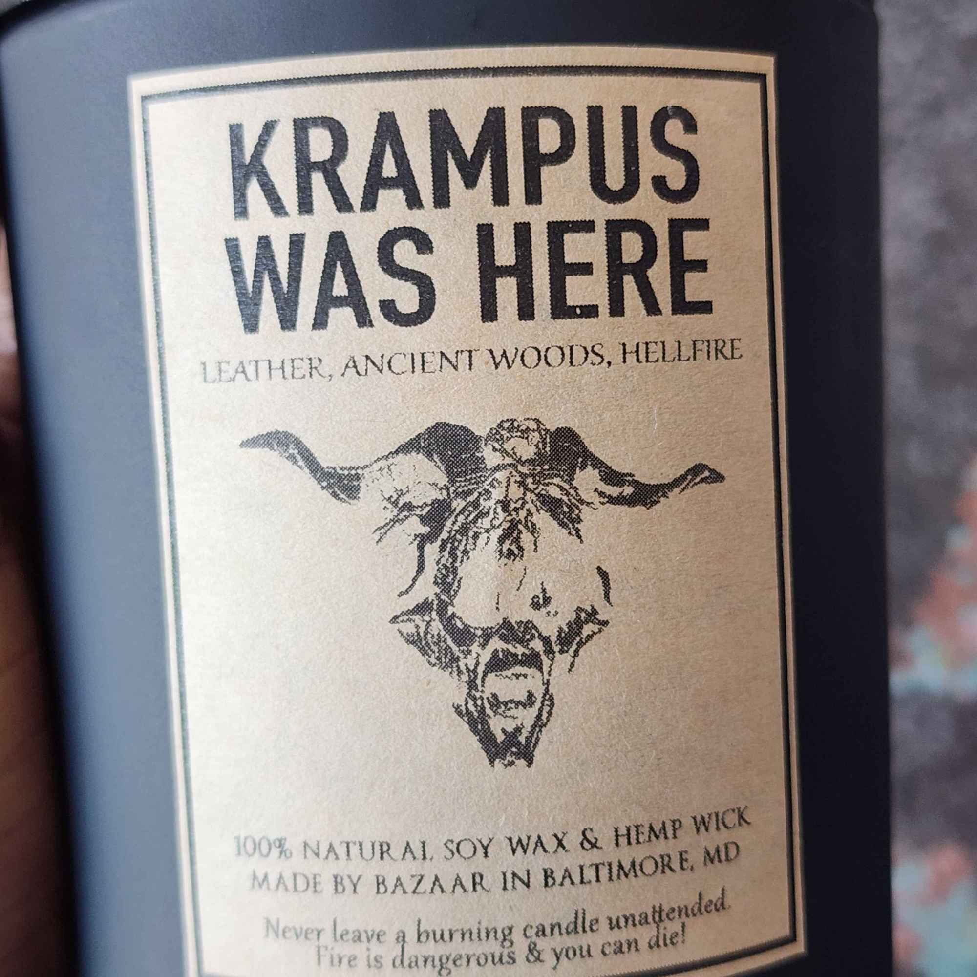 ID: a large candle from Bazaar in Baltimore in a matte black jar. the candle is called Krampus Was Here, and has an illustration of a goat head on the label. it smells like leather, ancient woods, and hellfire 