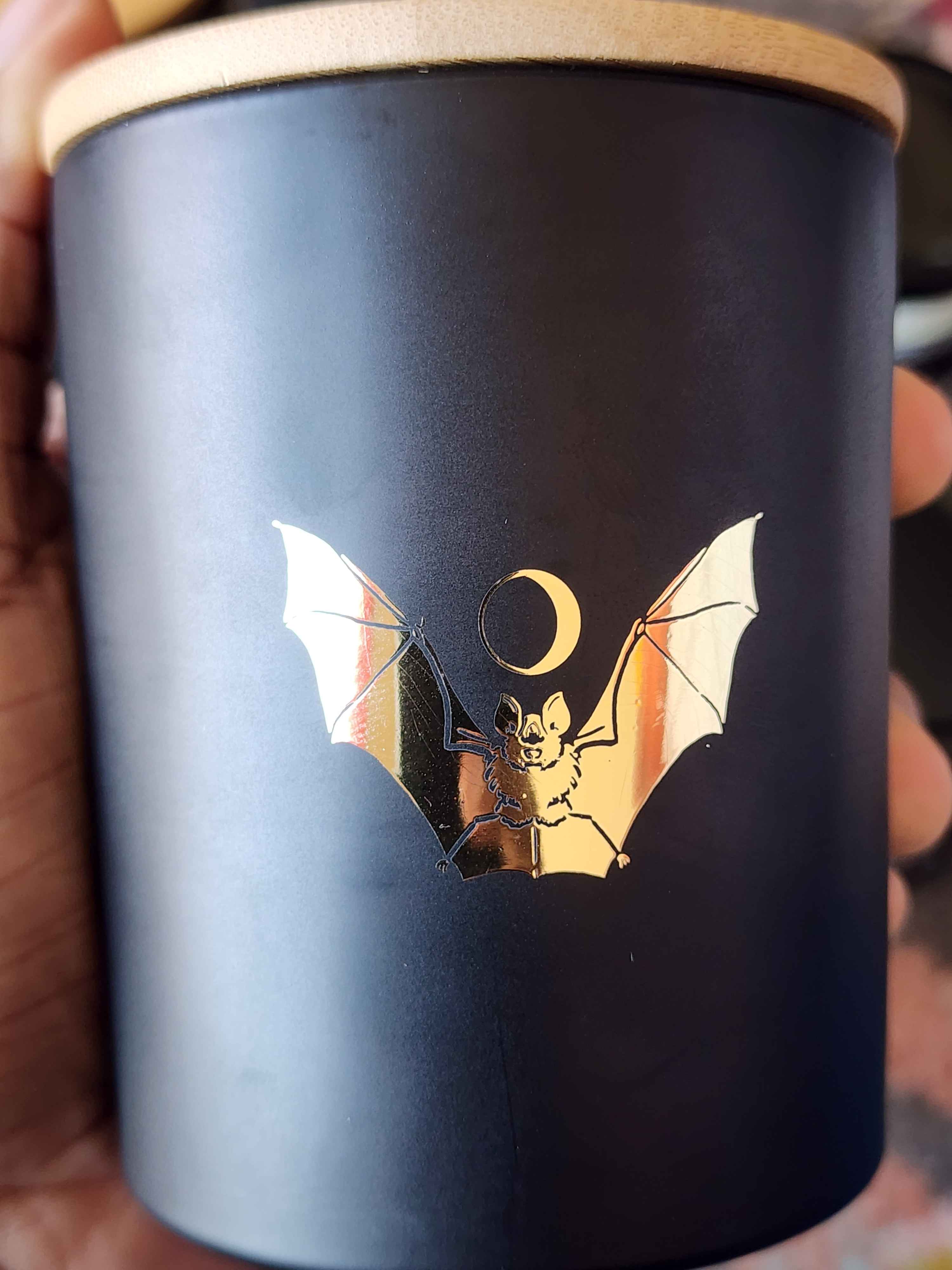 ID: a candle in a matte black jar from 13th Press called Midnight. a gold foil bat is spreads its wings across the front, and a crescent moon hovers over its head like a halo