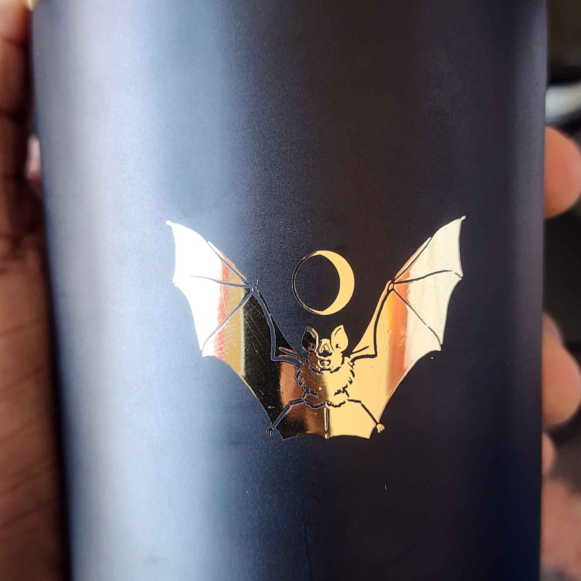 ID: a candle in a matte black jar from 13th Press called Midnight. a gold foil bat is spreads its wings across the front, and a crescent moon hovers over its head like a halo 