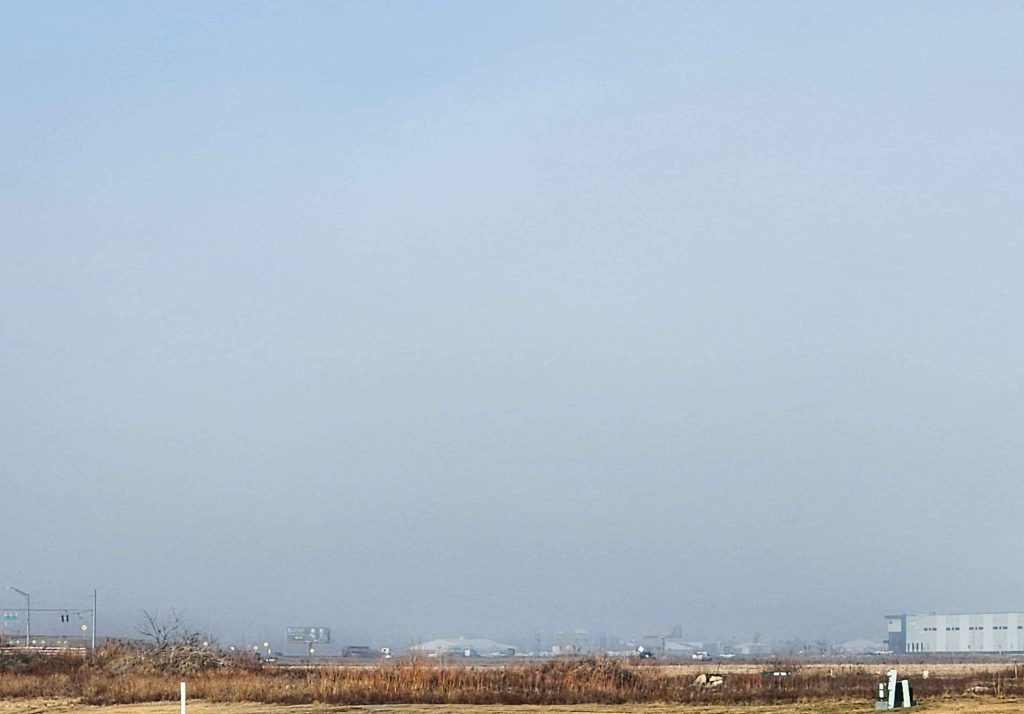 ID: a photo of a brown field and a highway in the distance. a wall of fog obscures most of a blue sky, and a warehouse lurks in the distance.