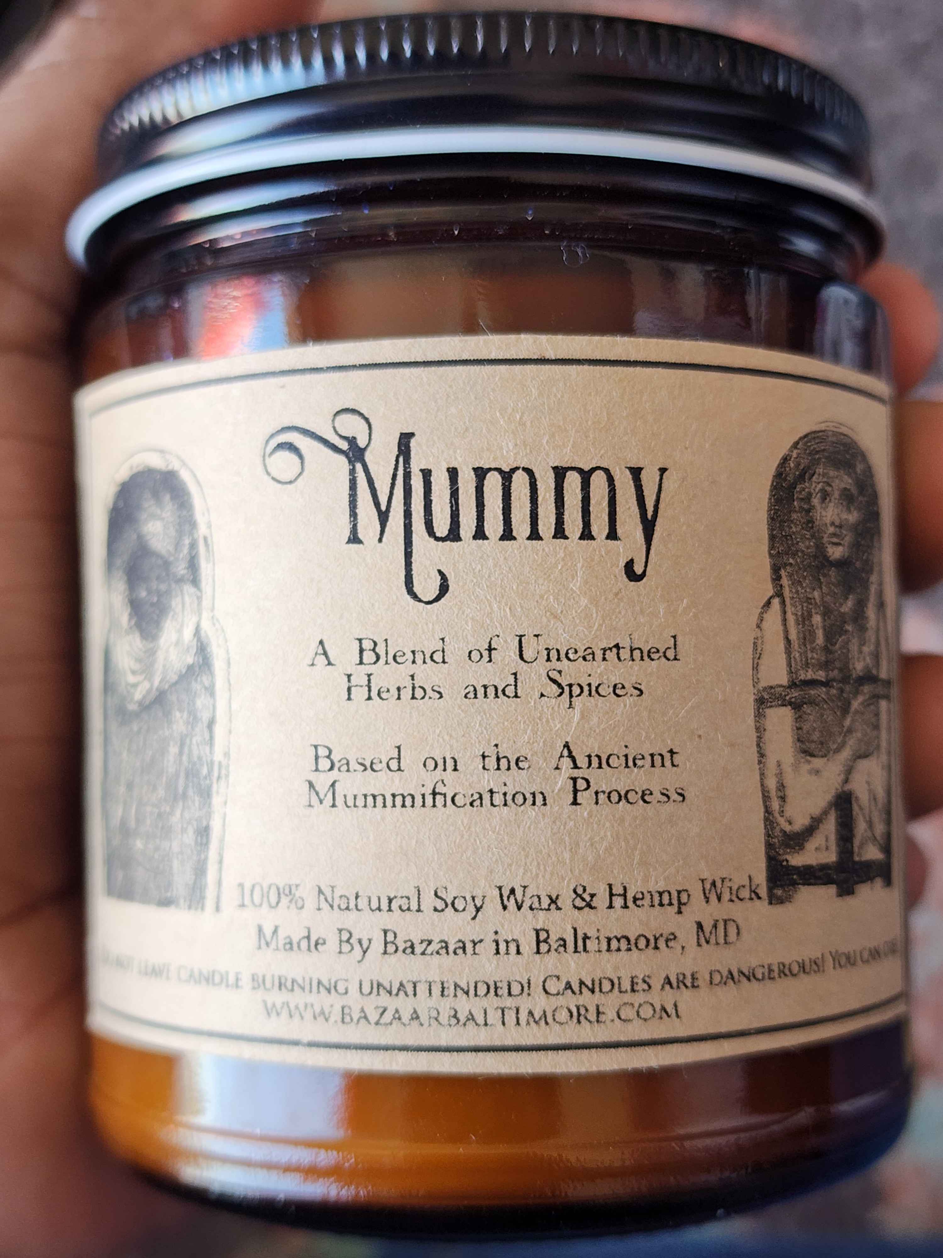 ID: an amber jar candle from Bazaar in Baltimore called Mummy. the label has illustrations of Egyptian style mummies and describes the candle as a blend of unearthed herbs and spices based on the ancient mummification process.