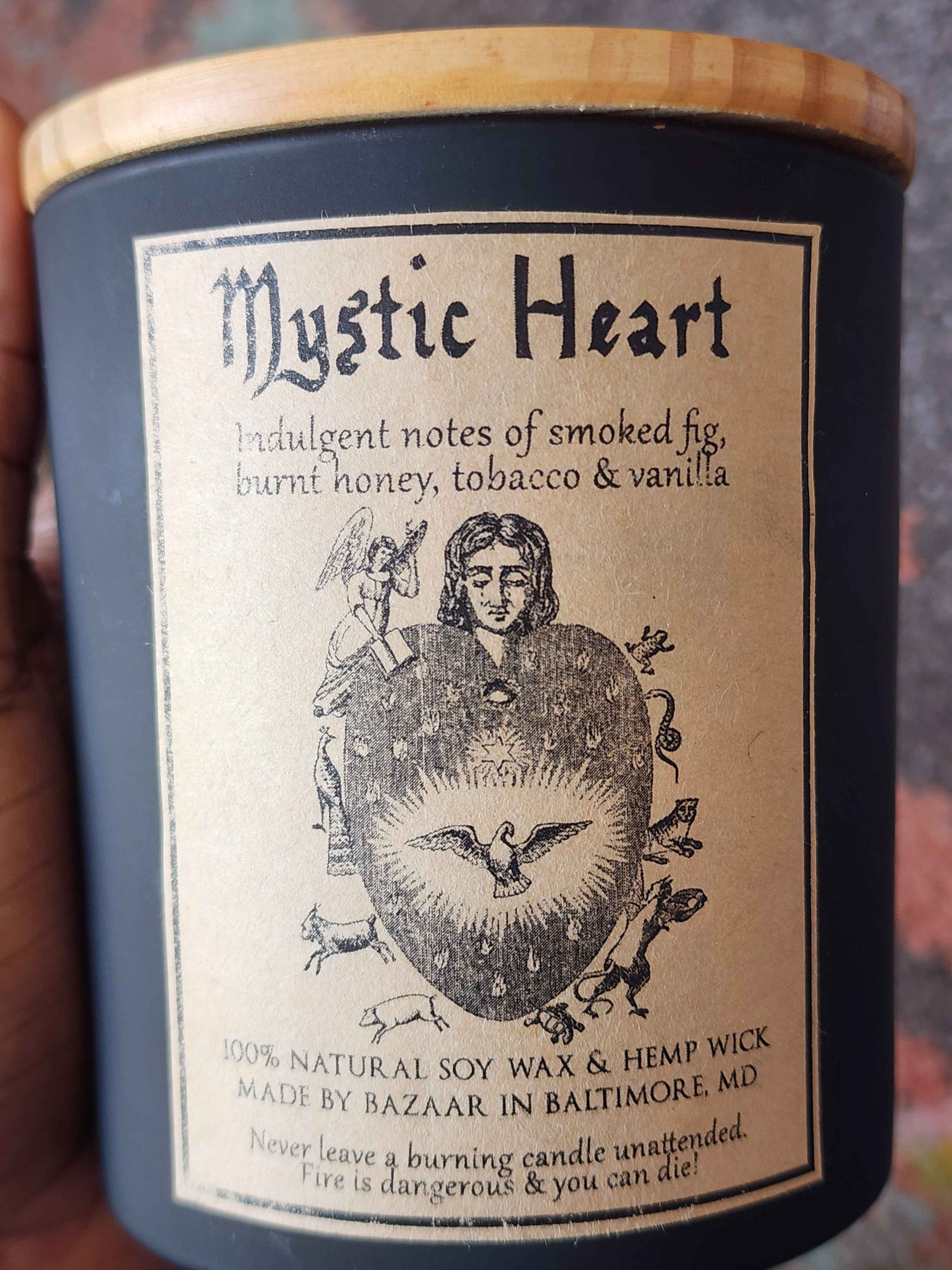 ID: a large candle from Bazaar in Baltimore in a matte black jar. the candle is called Mystic Heart. the label shows an intricate heart illustration with a dove in its center, woodland creatures around its borders, an angel on the top left, and a woman's head in the center. the candle has indulgent notes of smoked fig, burnt honey, tobacco, and vanilla