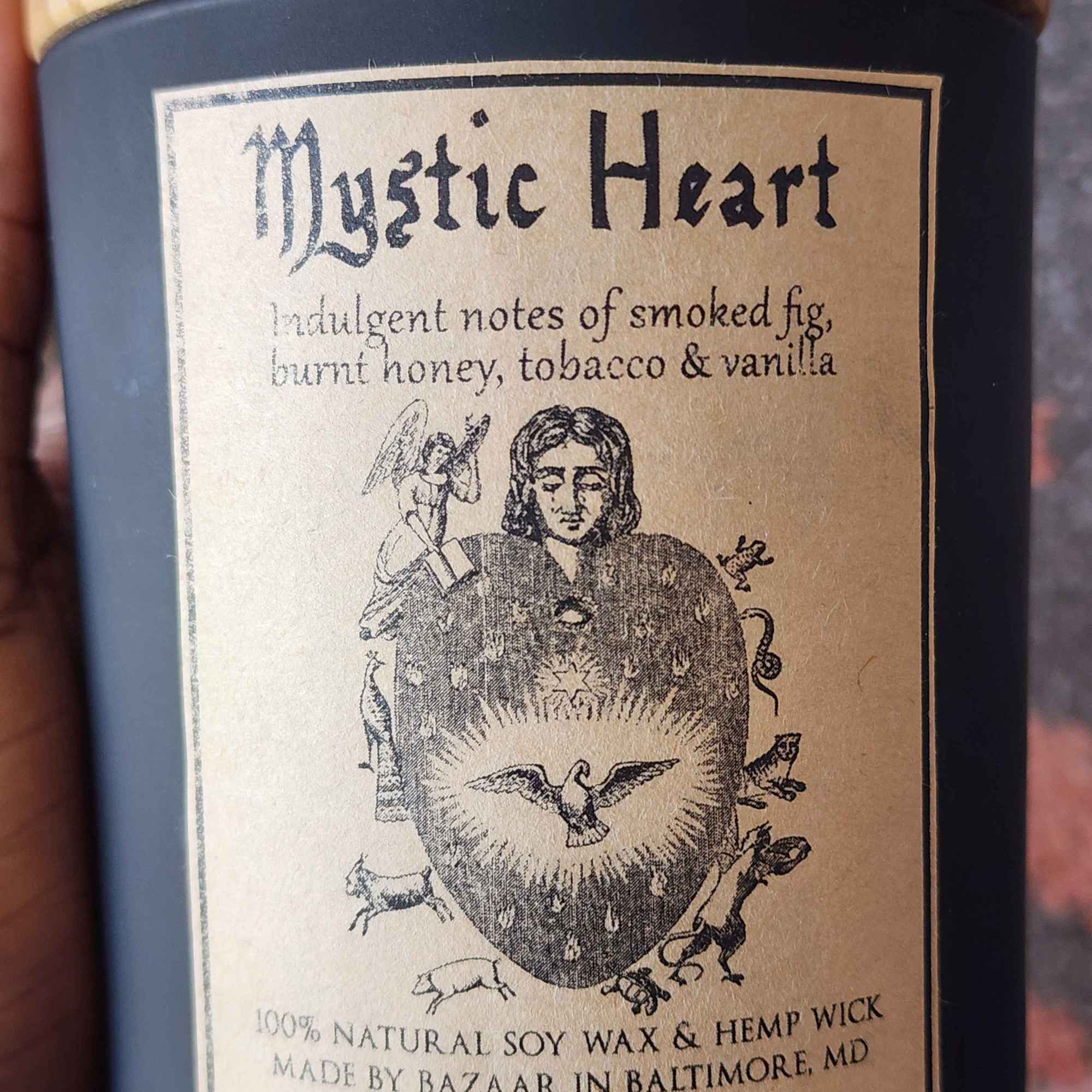 ID: a large candle from Bazaar in Baltimore in a matte black jar. the candle is called Mystic Heart. the label shows an intricate heart illustration with a dove in its center, woodland creatures around its borders, an angel on the top left, and a woman's head in the center. the candle has indulgent notes of smoked fig, burnt honey, tobacco, and vanilla