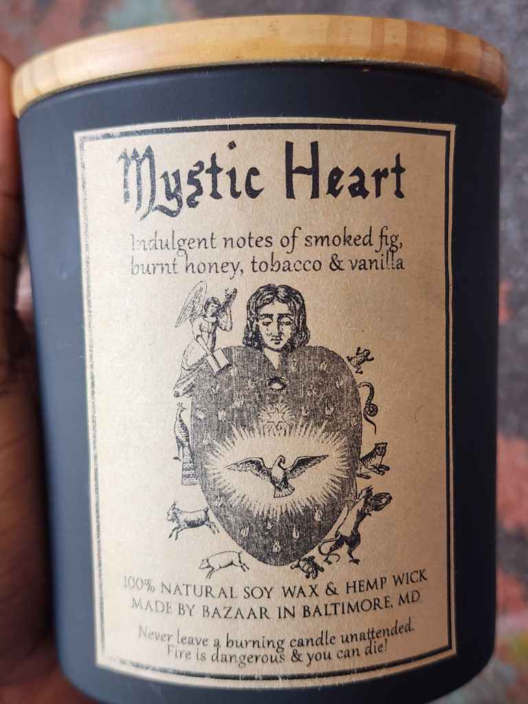 ID: a large candle from Bazaar in Baltimore in a matte black jar. the candle is called Mystic Heart. the label shows an intricate heart illustration with a dove in its center, woodland creatures around its borders, an angel on the top left, and a woman's head in the center. the candle has indulgent notes of smoked fig, burnt honey, tobacco, and vanilla