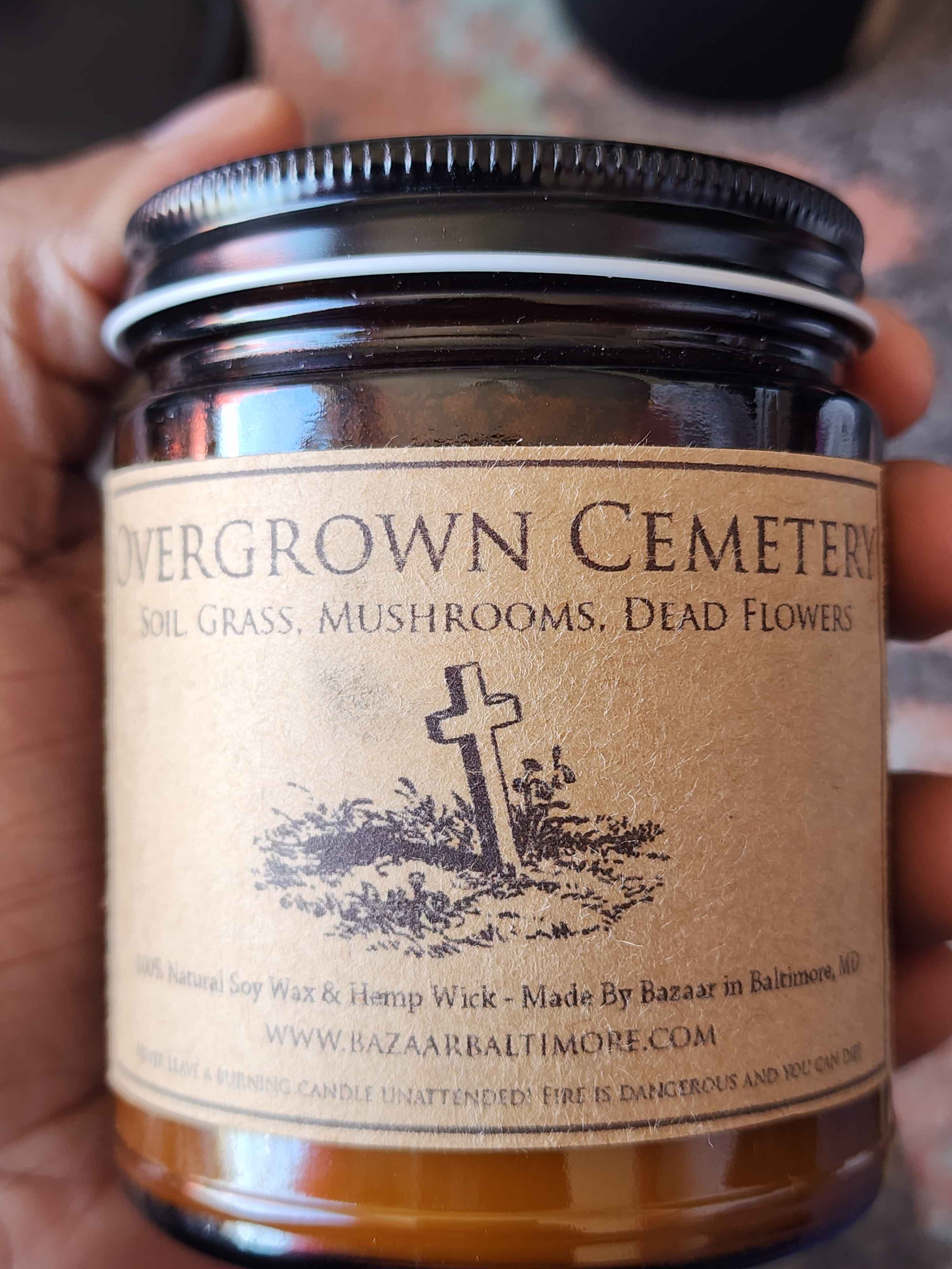 ID: an amber jar candle from Bazaar in Baltimore called Overgrown Cemetery. a small illustration of a cemetery hill with a cross is on the label. the candle has notes of soil, grass, mushrooms, and dead flowers