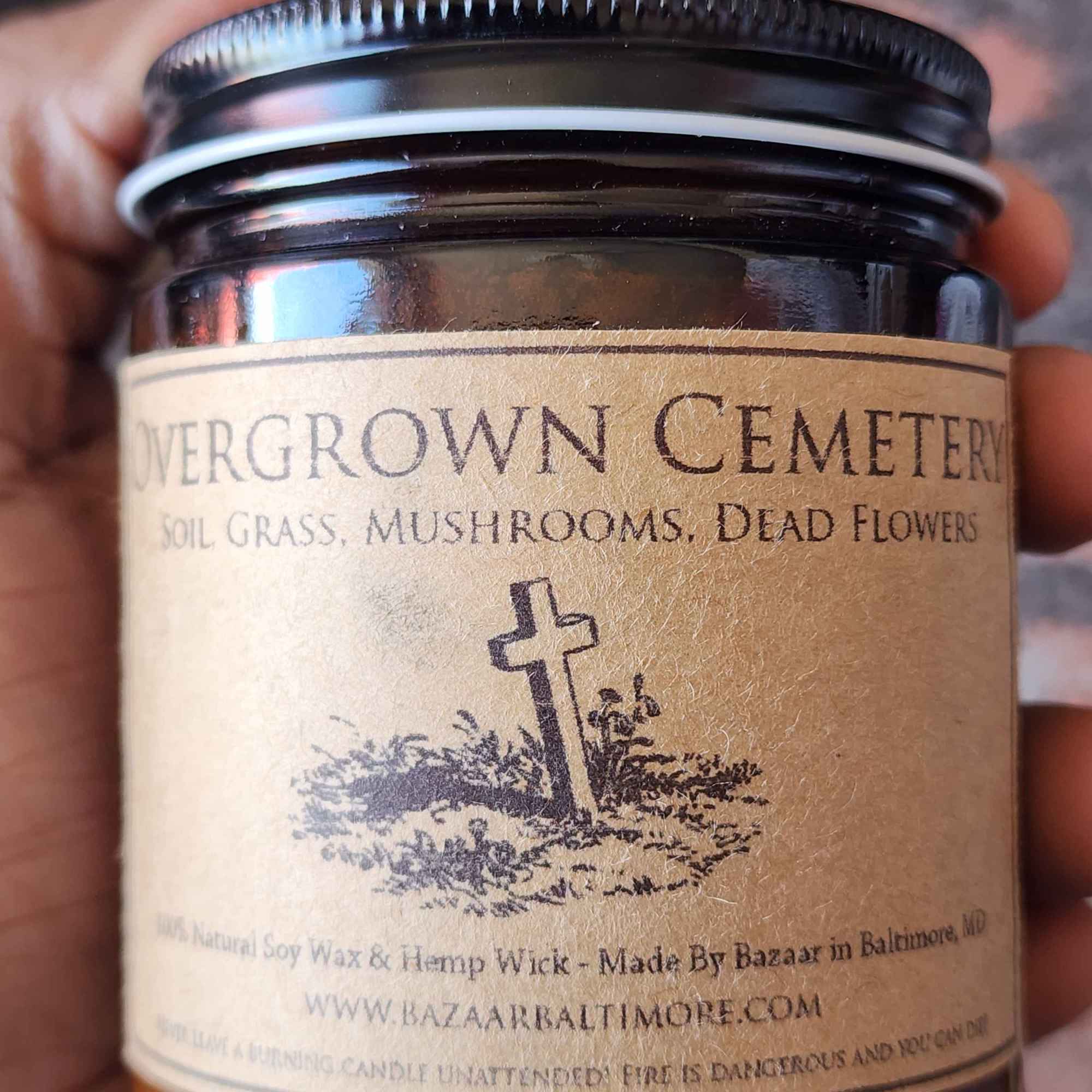 ID: an amber jar candle from Bazaar in Baltimore called Overgrown Cemetery. a small illustration of a cemetery hill with a cross is on the label. the candle has notes of soil, grass, mushrooms, and dead flowers