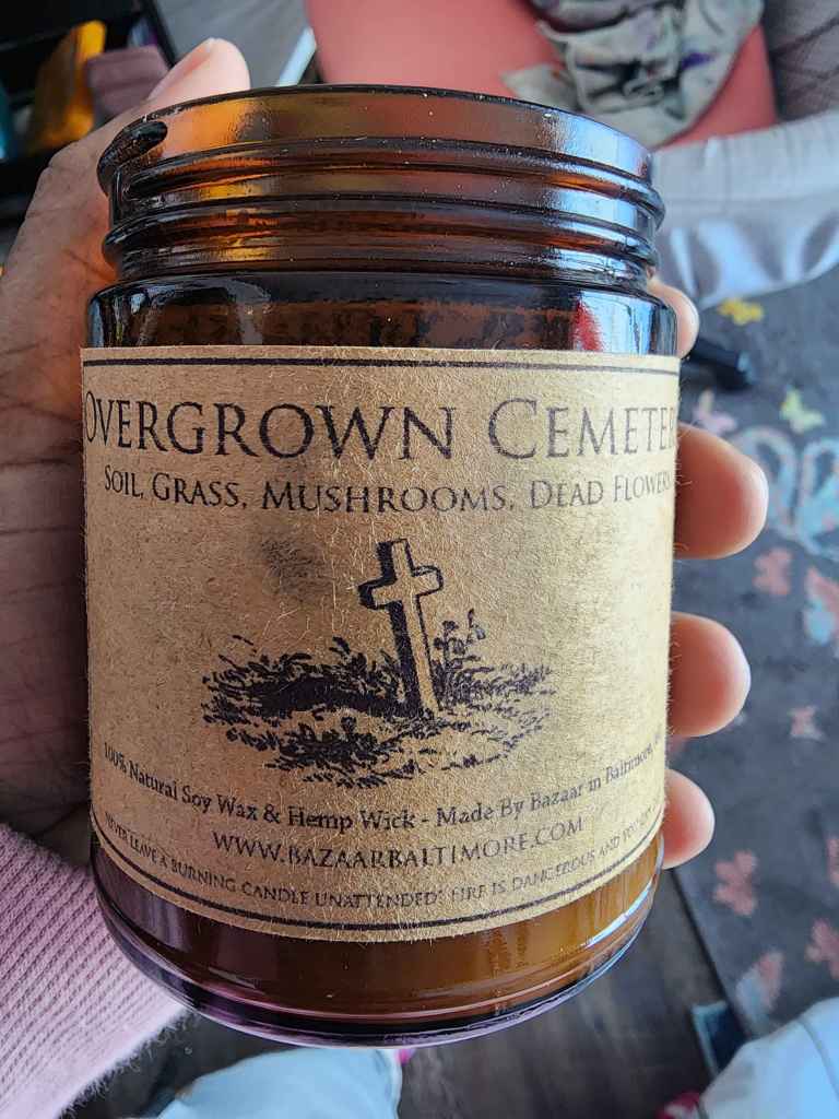 ID: a candle from Bazaar in Baltimore in an amber glass jar labeled "Overgrown Cemetery" with notes of soil, grass, mushrooms, and dead flowers.