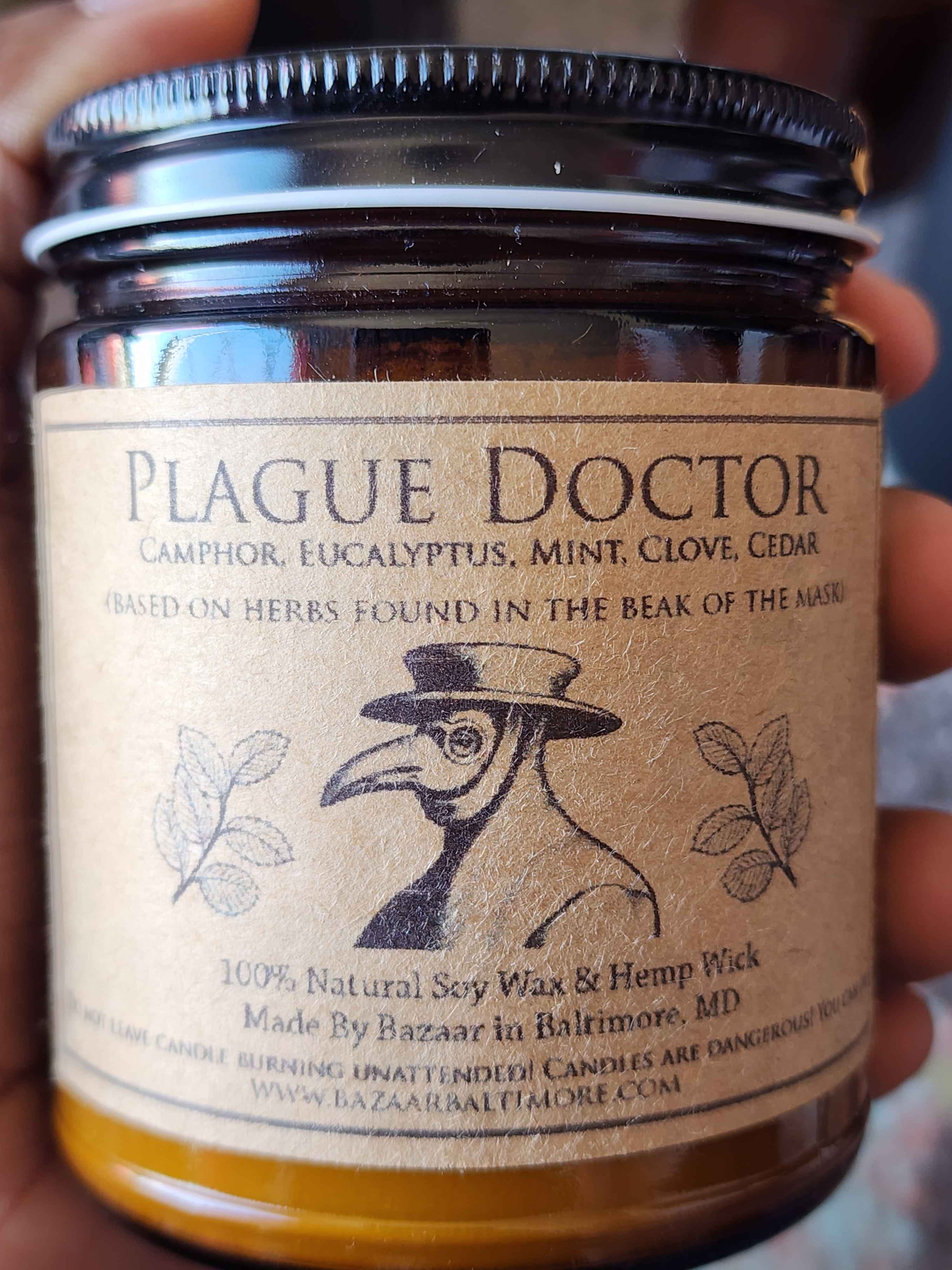 ID: an amber jar candle from Bazaar in Baltimore called Plague Doctor. the label shows an illustration of a figure in a top hat and an old bird's mask. the candle has notes of camphor, eucalyptus, mint, clove, and cedar. the scent is based on herbs found in the beak of similar masks used by doctors of that era.