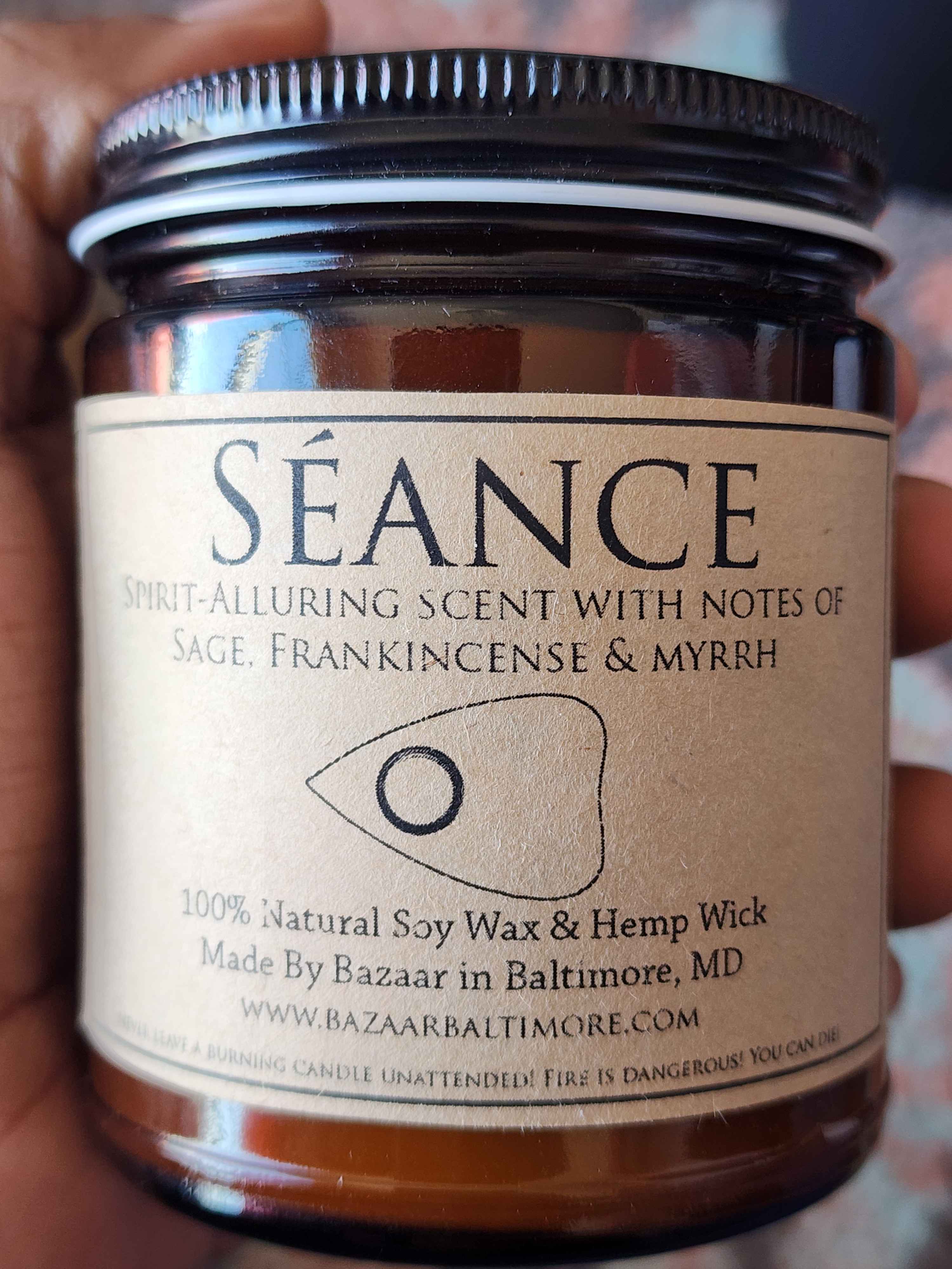 ID: an amber jar candle from Bazaar in Baltimore called Seance, an illustration of a ouija board piece is on the label. the candle is described as a spirit-alluring scent with notes of sage, frankincense, and myrrh