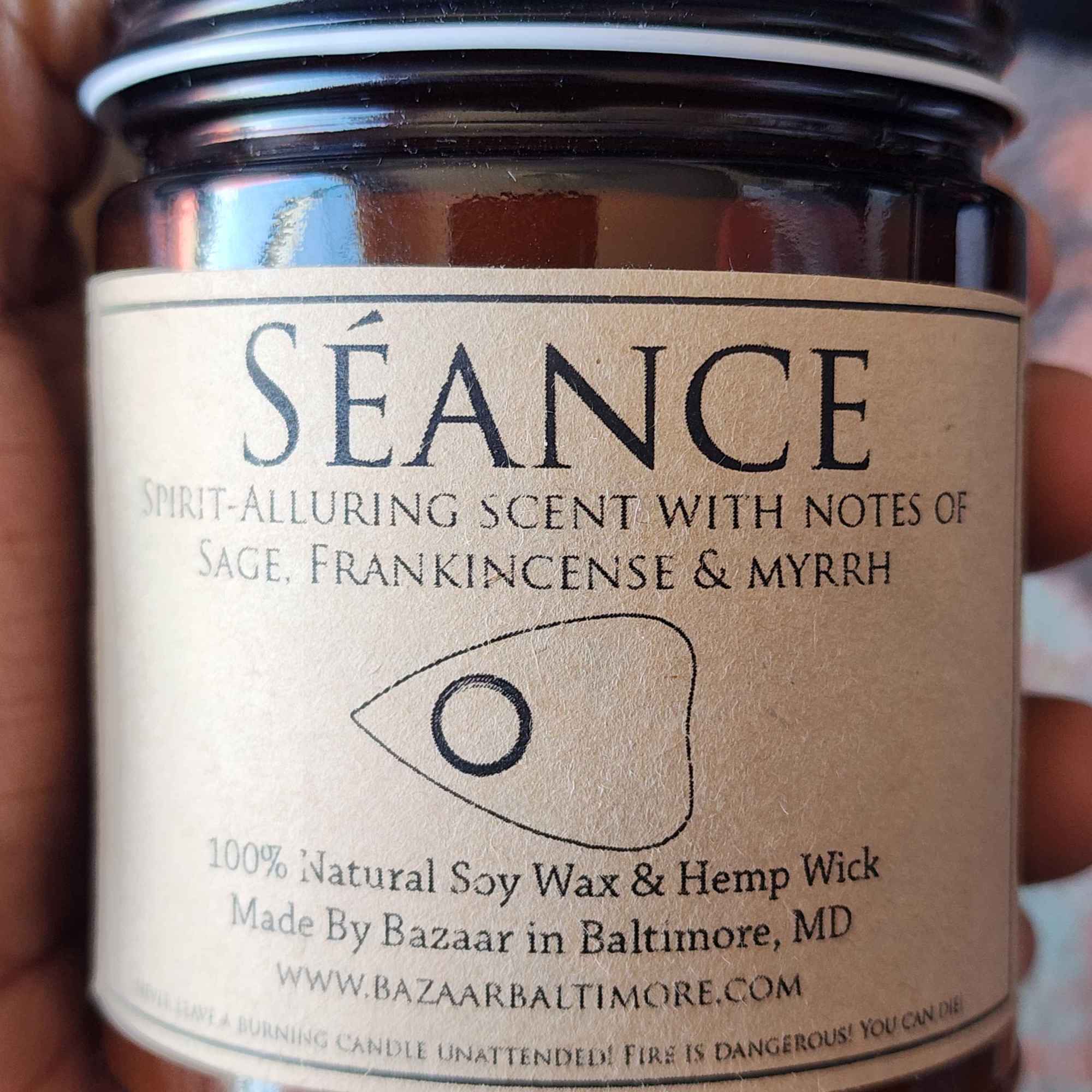 ID: an amber jar candle from Bazaar in Baltimore called Seance, an illustration of a ouija board piece is on the label. the candle is described as a spirit-alluring scent with notes of sage, frankincense, and myrrh